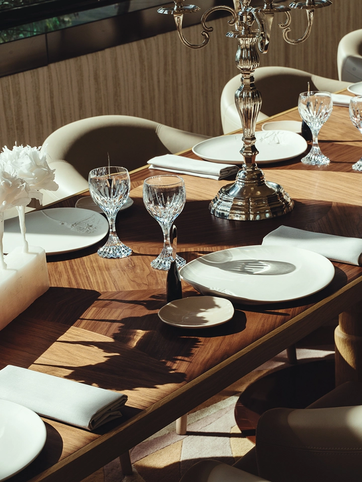 Table en bois élégamment dressée avec assiettes blanches, verres en cristal, serviettes pliées, bouquet de fleurs blanches et chandelier en métal argenté.
