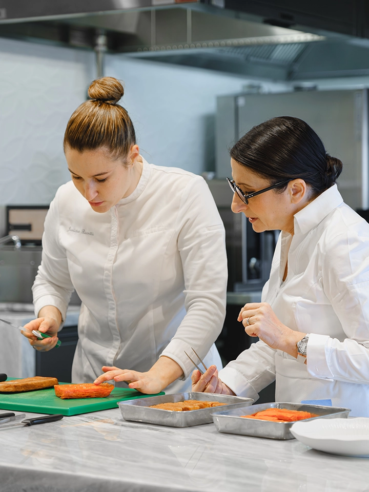 Deux cuisinières en blouse blanche préparent et examinent des aliments dans une cuisine professionnelle.