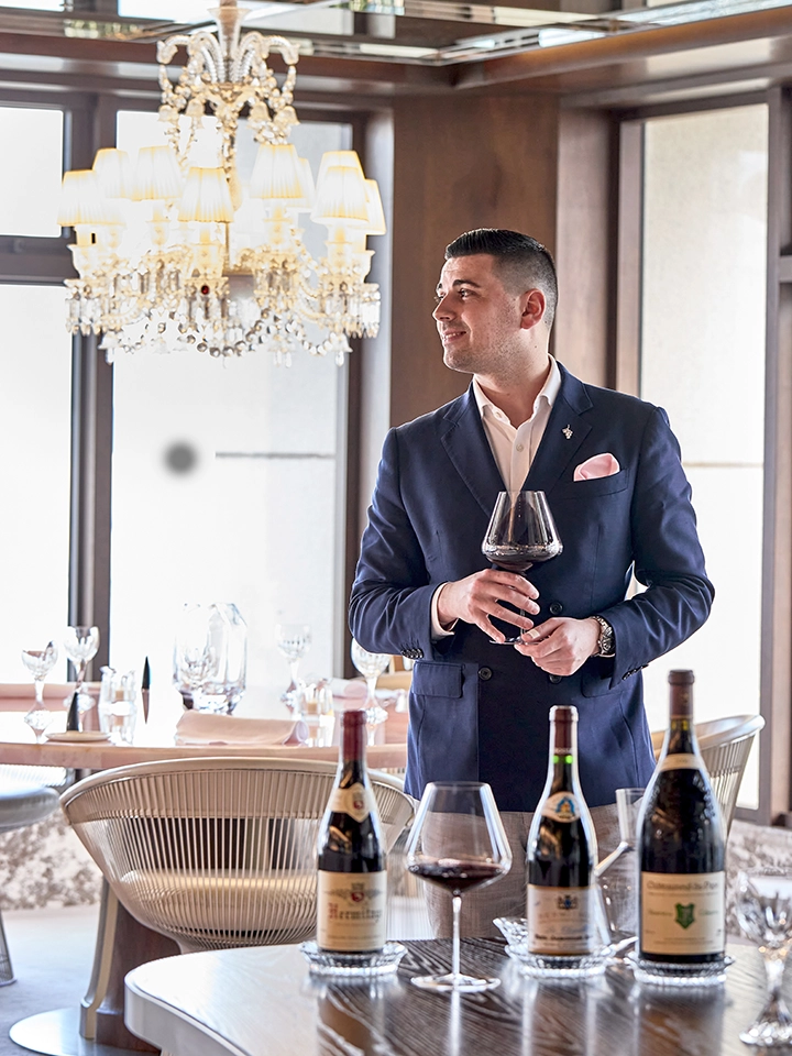 Un sommelier en costume bleu tenant un verre de vin rouge dans une salle élégante avec des bouteilles de vin sur la table devant lui.