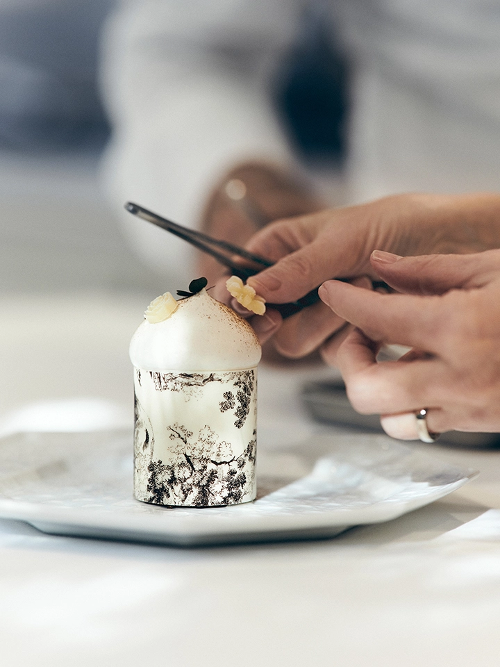 Dessert élégant composé d’une mousse blanche décorée de petites fleurs, présenté sur une assiette, mains ajustant la finition.