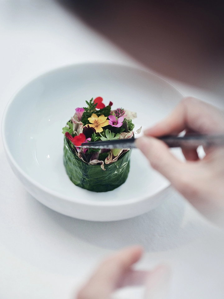 Plat artistique composé de fleurs comestibles colorées enroulées dans une feuille verte, présenté dans un bol blanc, main tenant des baguettes prête à déguster.