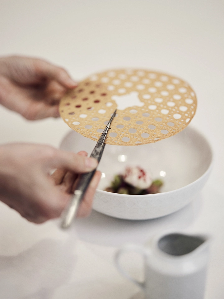 Main tenant une fine galette ronde ajourée, placée au-dessus d’un bol blanc contenant un dessert.