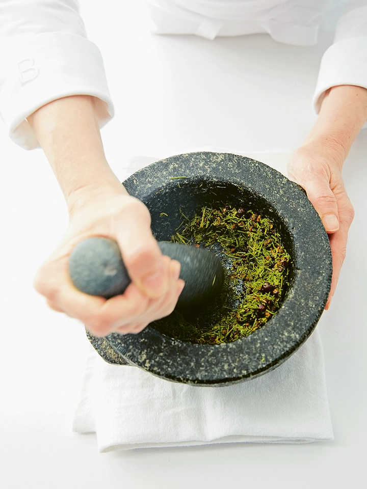 Deux mains utilisent un mortier en pierre noire pour broyer des herbes vertes à l’aide d’un pilon.