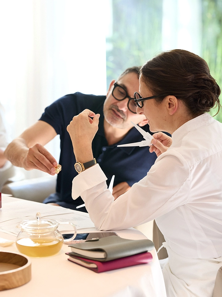 Deux personnes discutant en examinant un échantillon de parfum avec des mouillettes, assises à une table avec un thé en pot en verre.