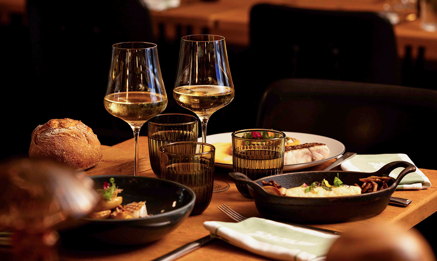 Table en bois avec deux verres de vin blanc, des verres à eau, un pain rond, et plusieurs plats gastronomiques servis dans une ambiance chaleureuse de restaurant.
