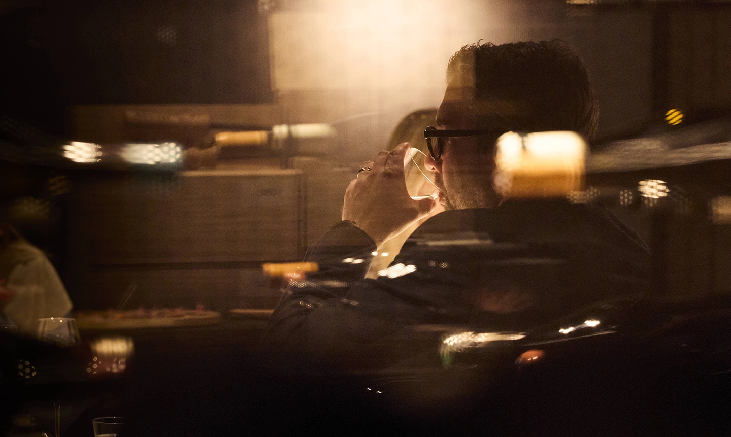 Homme portant des lunettes buvant dans un verre dans un environnement sombre avec des reflets lumineux flous.