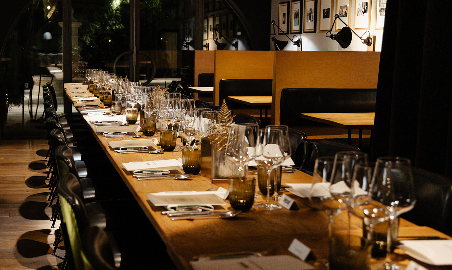 Table en bois longue dressée avec des verres à vin, des assiettes et des couverts dans un restaurant élégant, avec des sièges en cuir noir et un décor moderne.