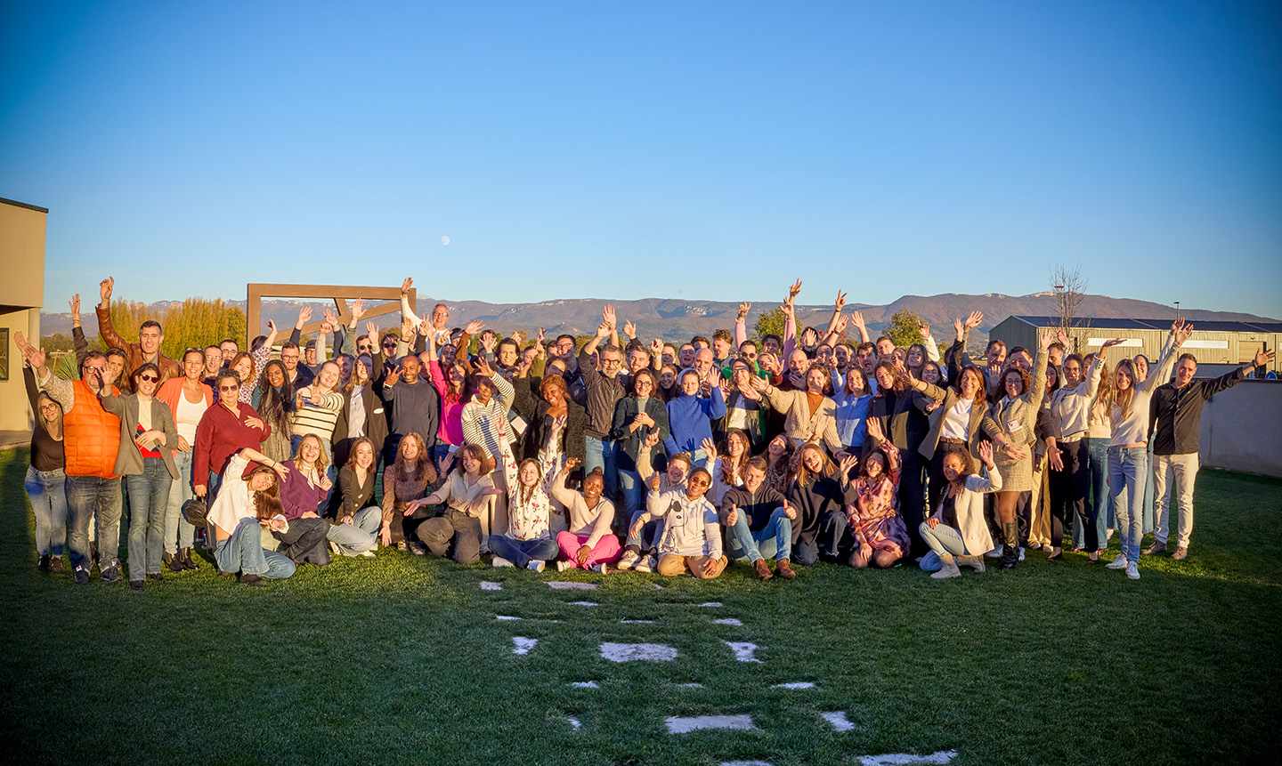 Groupe divers de personnes souriantes posant ensemble en plein air sur une pelouse avec montagnes en arrière-plan, sous un ciel bleu clair.