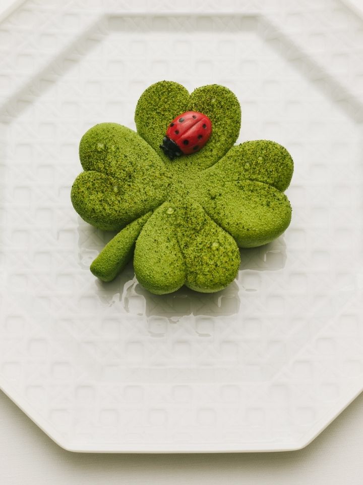 Dessert vert en forme de trèfle avec une coccinelle rouge dessus, posé sur une assiette blanche texturée.