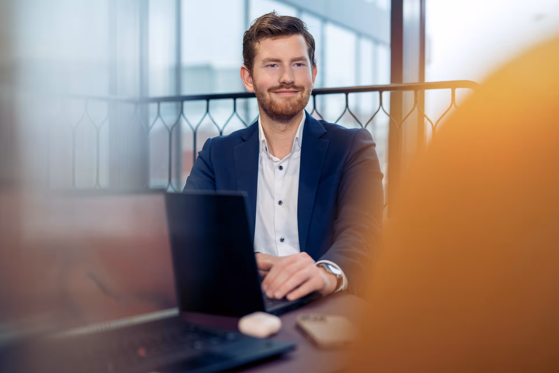 Smiling man in a navy blazer working on a laptop in a modern office with large windows.