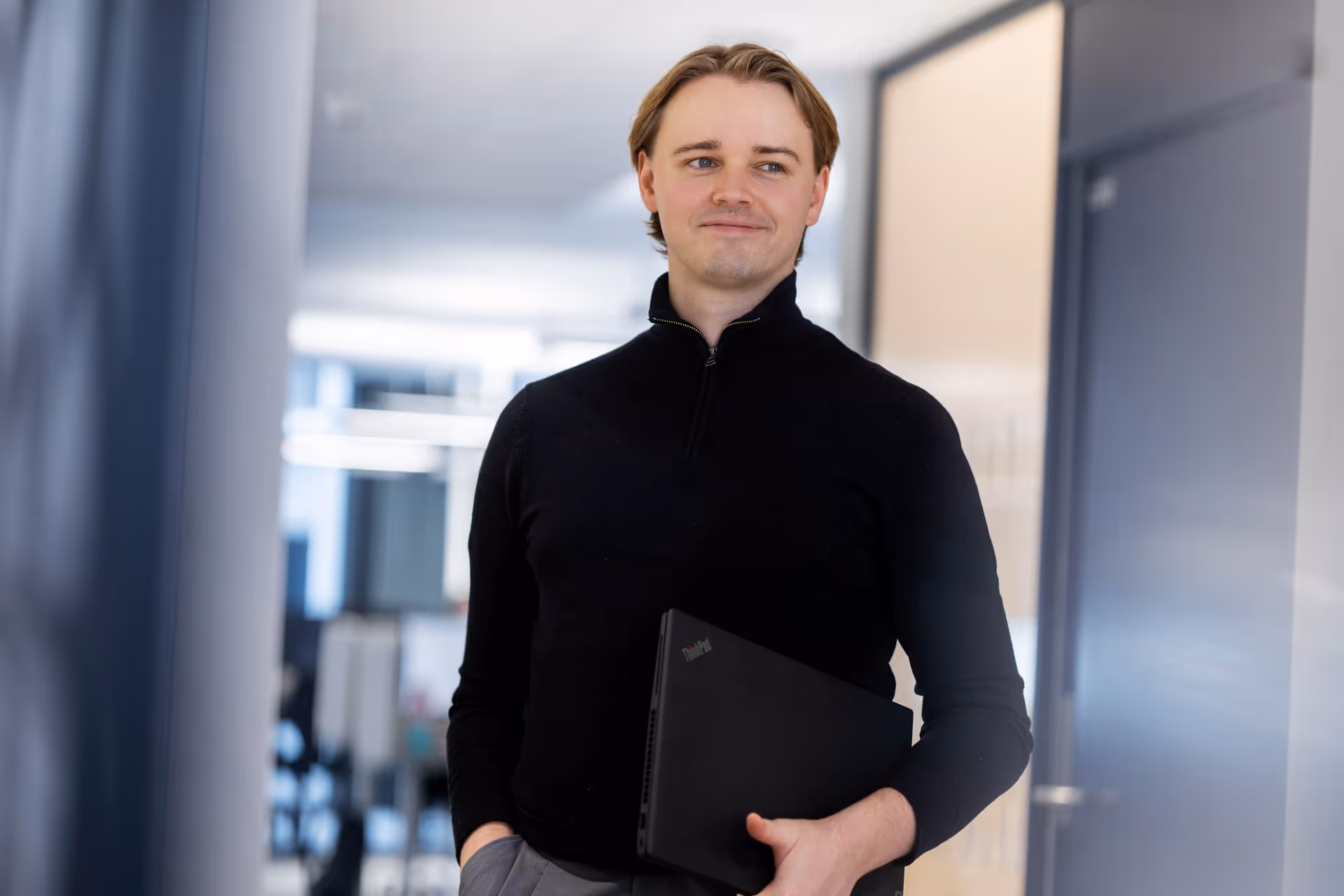 Young man in black sweater holding a closed laptop in an office hallway.