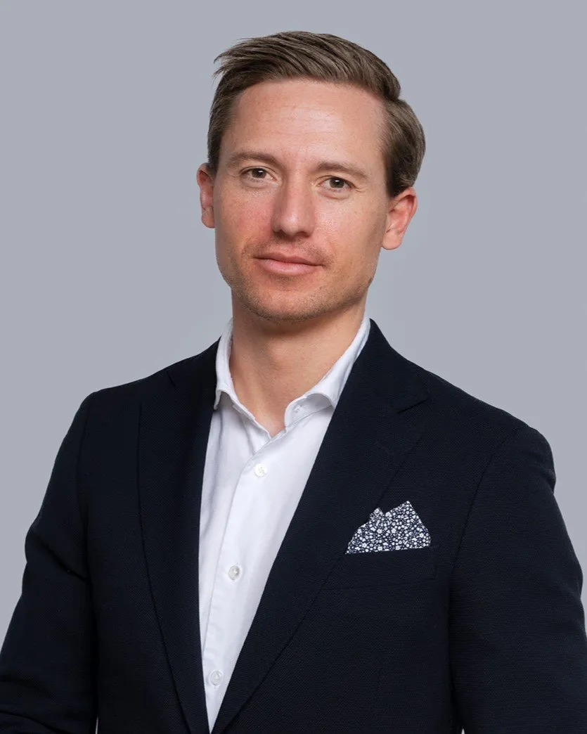 Man wearing a dark suit jacket with a white shirt and patterned pocket square against a plain gray background.