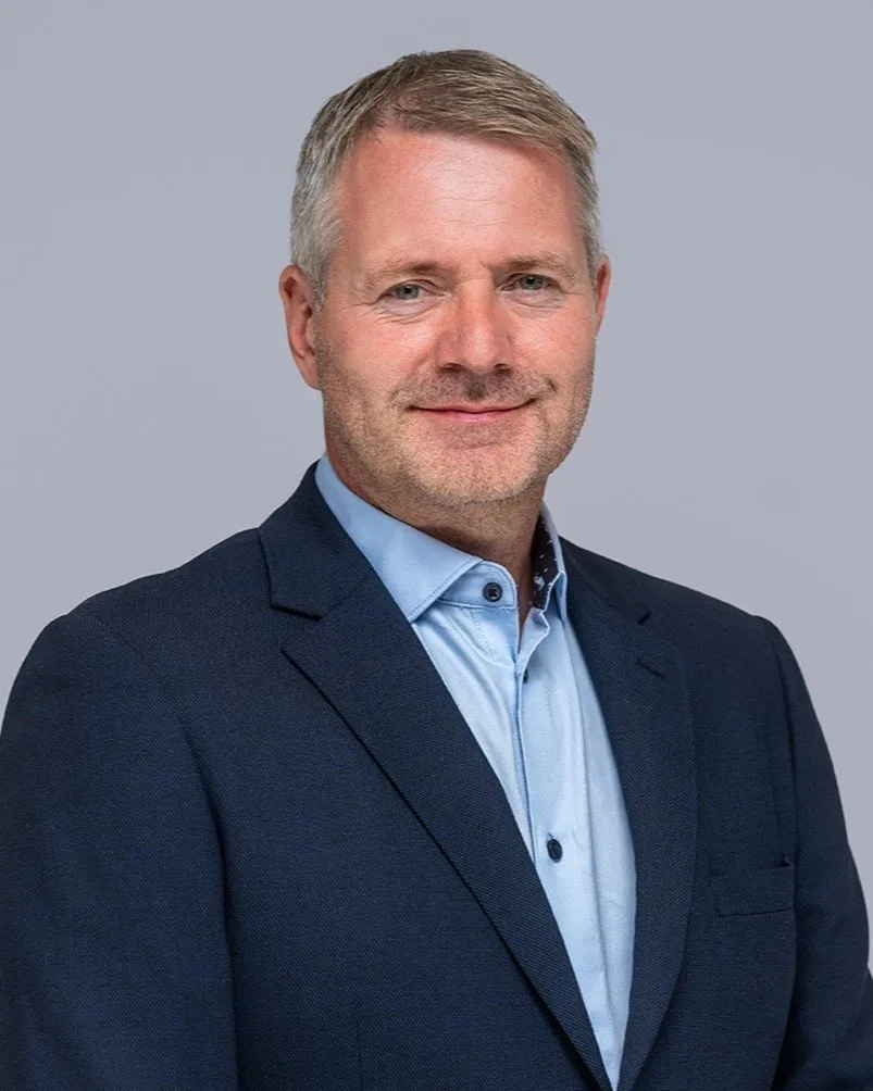 Smiling middle-aged man with short gray hair wearing a blue dress shirt and dark blazer against a plain background.