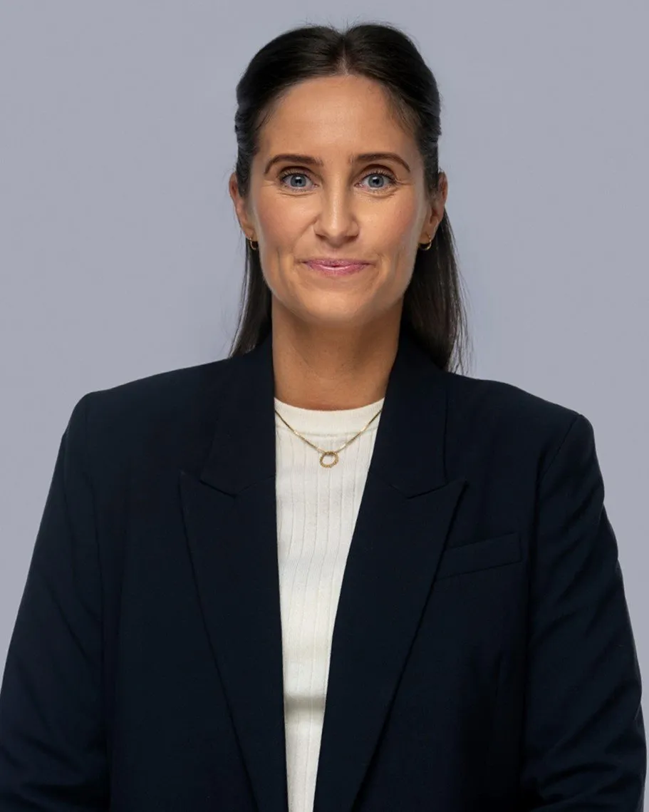 Smiling woman with dark hair wearing a black blazer, white top, and gold necklace on a gray background.