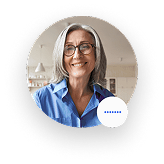 Smiling middle-aged woman with gray hair and glasses wearing a blue shirt in a home setting.