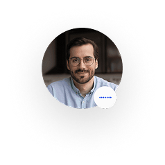 Smiling man with glasses and beard wearing a light blue shirt in an indoor setting.