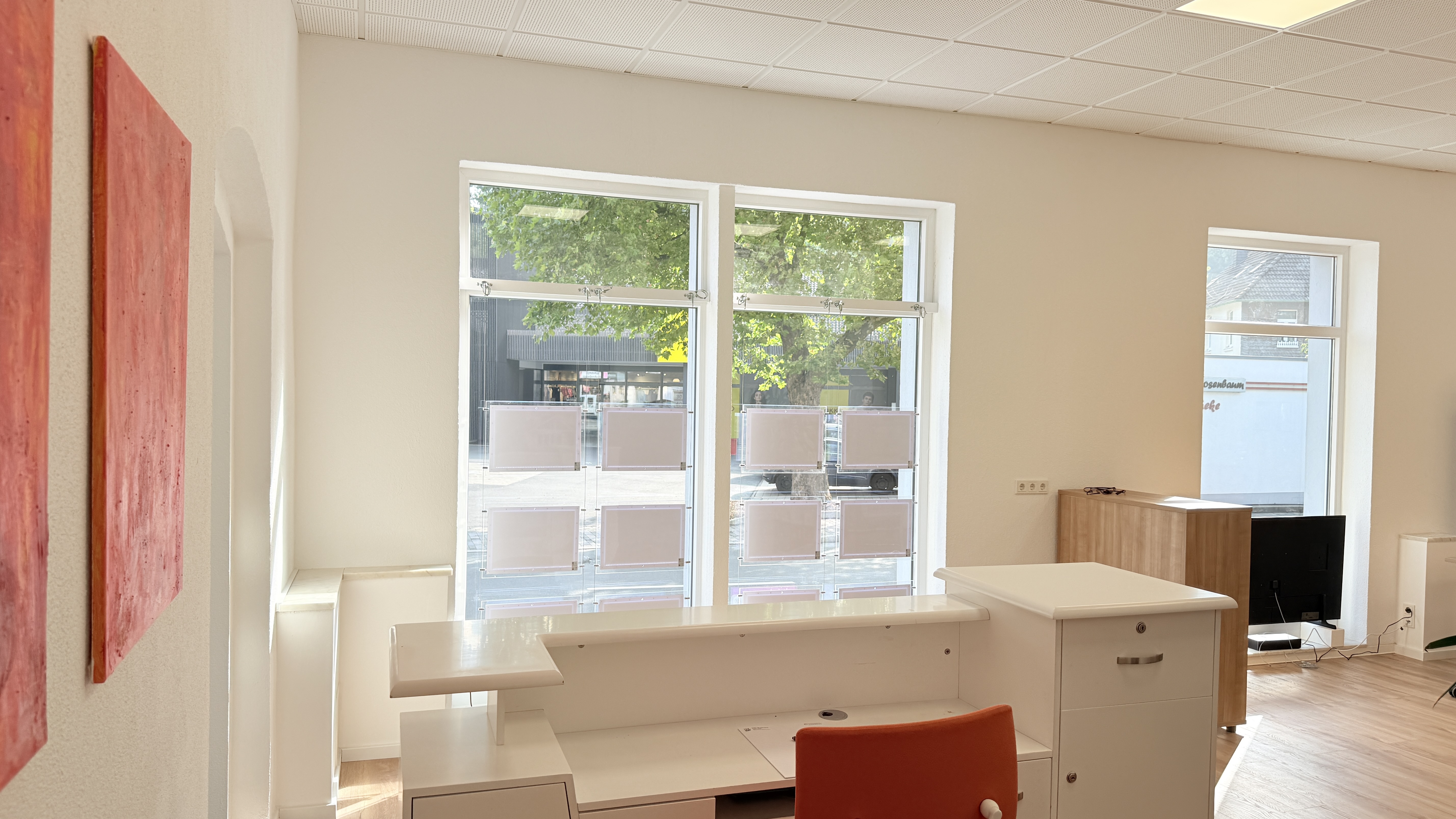 Bright modern office interior with white desk, orange chair, large windows, and minimal wooden furniture.
