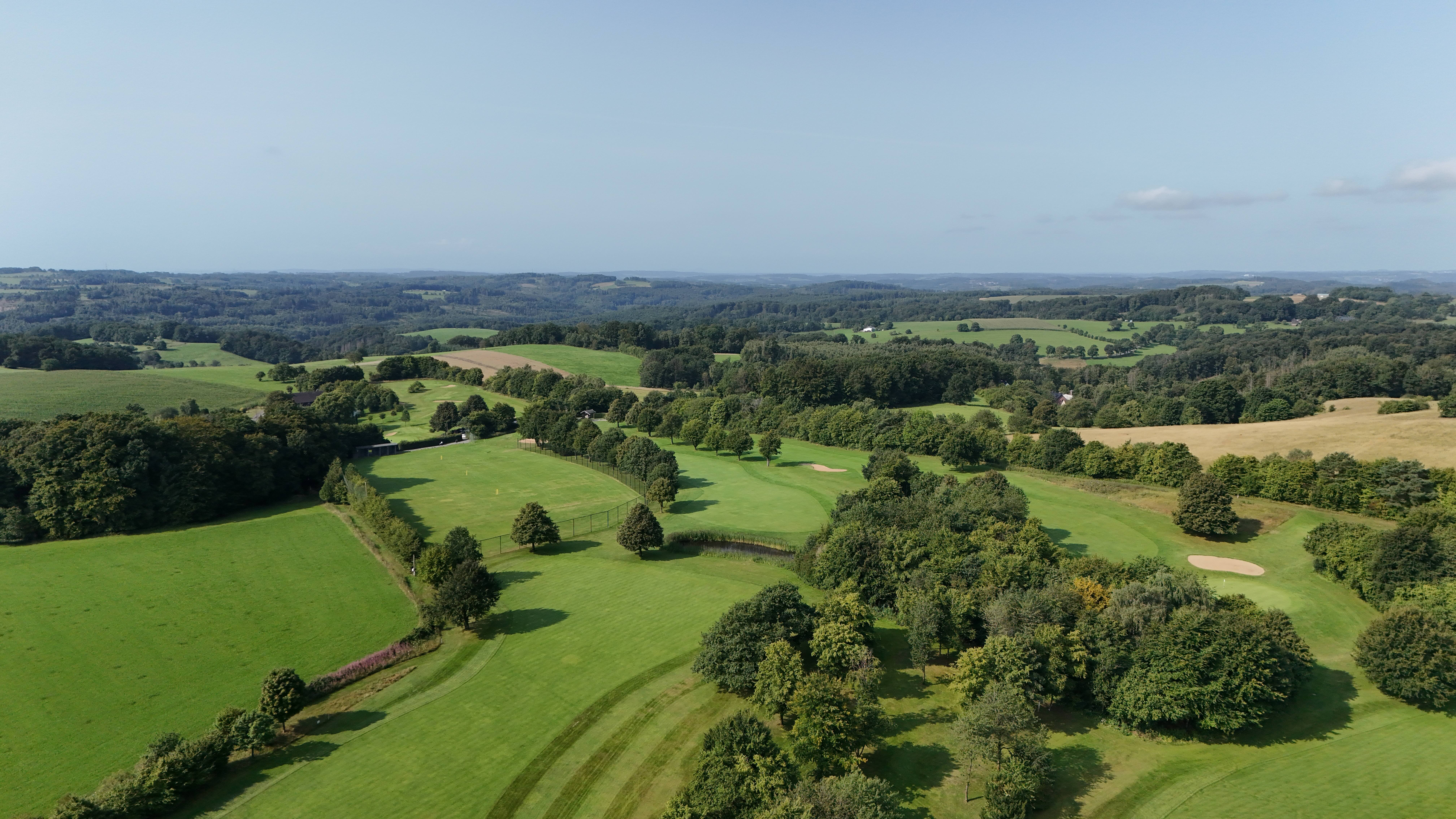 Aerial view of a golf course with lush green fairways, sand bunkers, trees, and surrounding rolling hills under a clear blue sky.