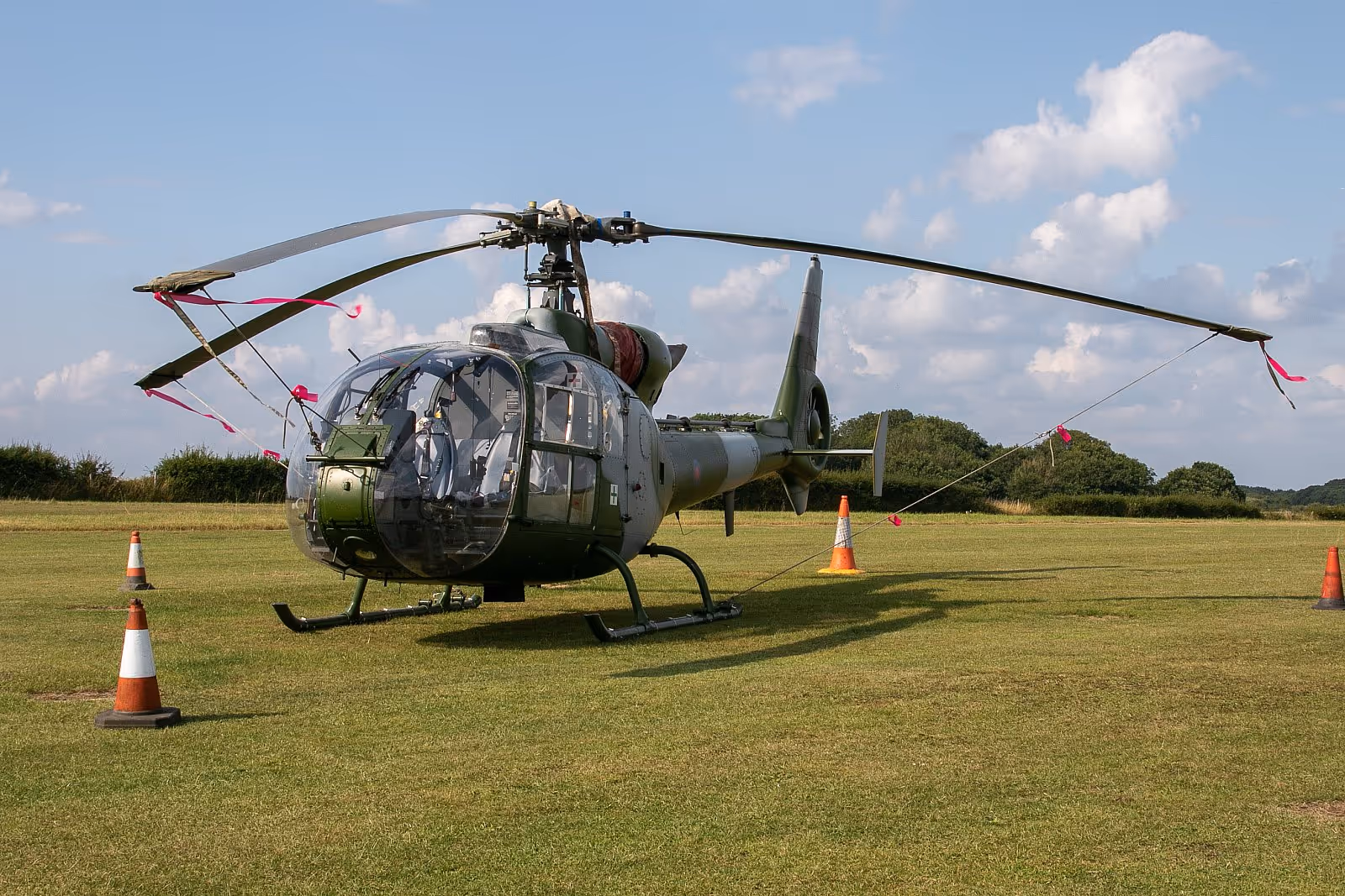 A military helicopter is parked on a grass field, secured with ropes and orange cones.
