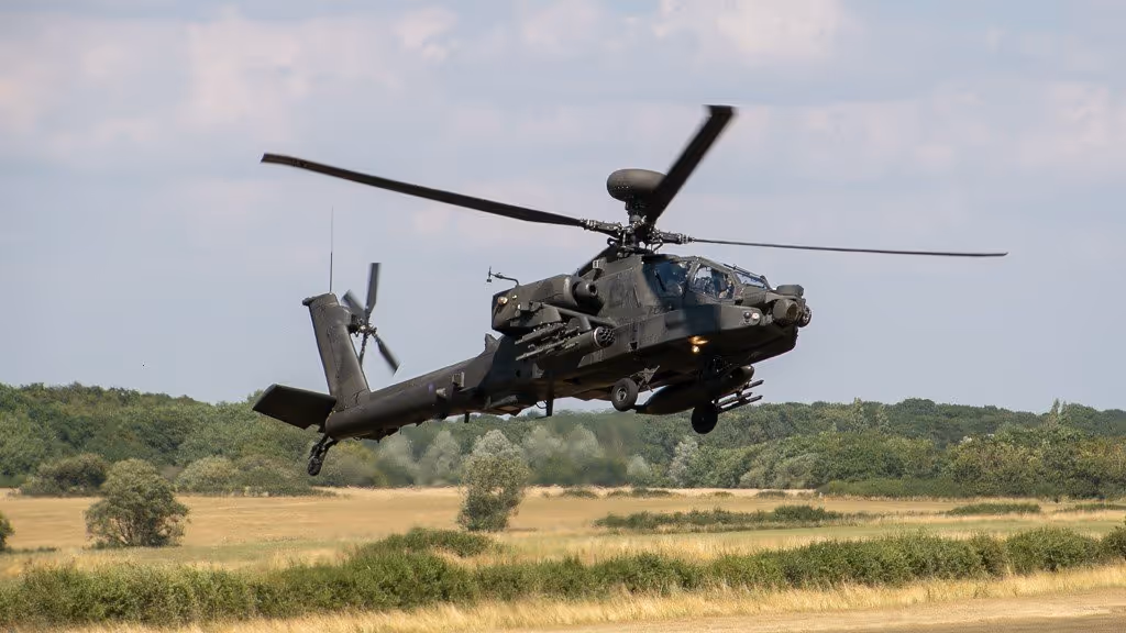 A military helicopter hovers low above a field, with trees visible in the background.