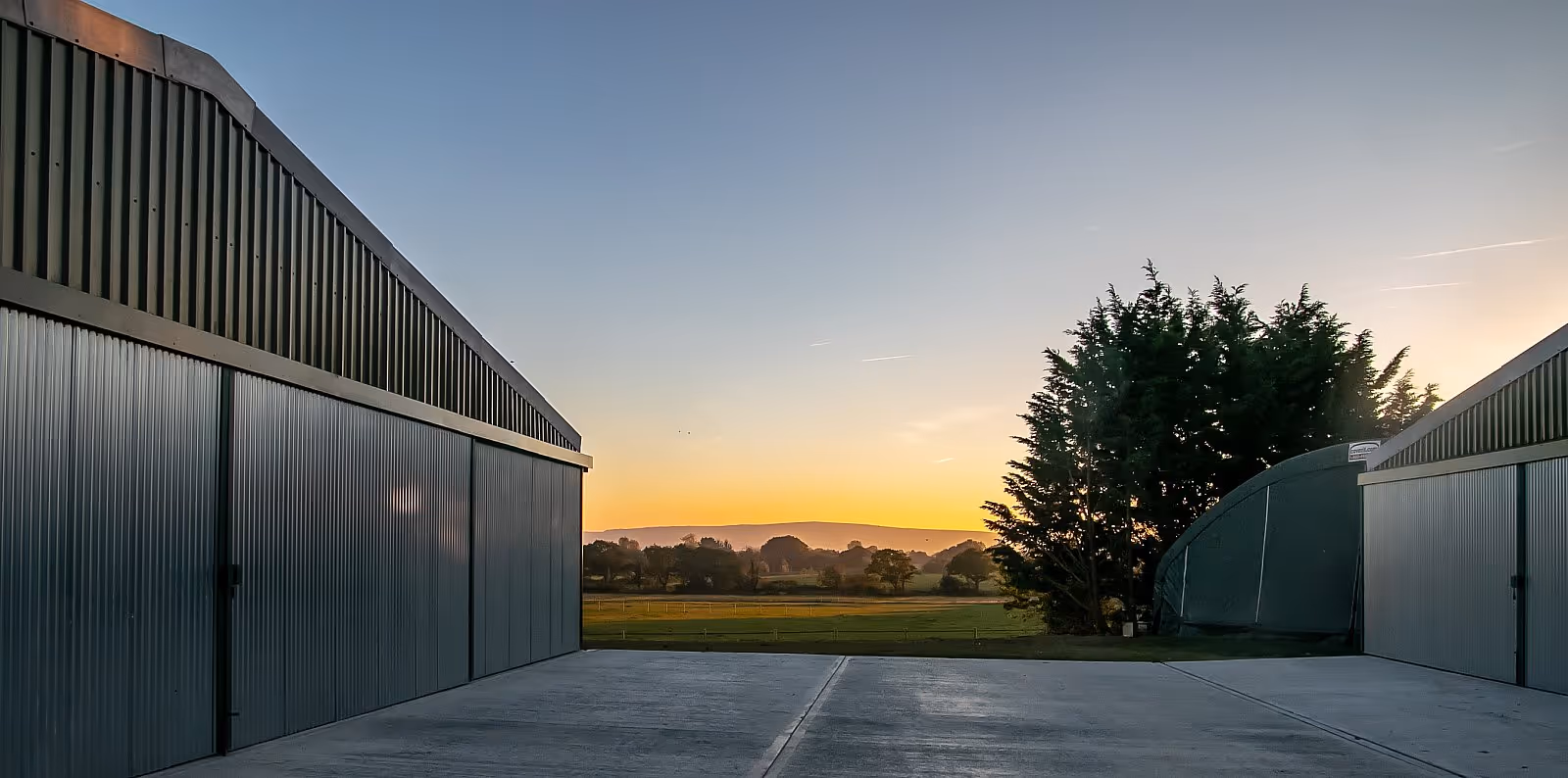 Industrial buildings with large metal doors at sunset, overlooking distant hills and a tree.