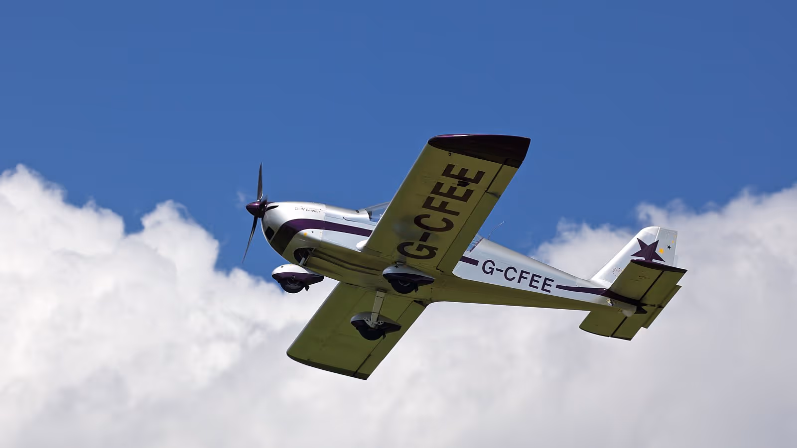 A small propeller airplane flies through a clear blue sky with white clouds.