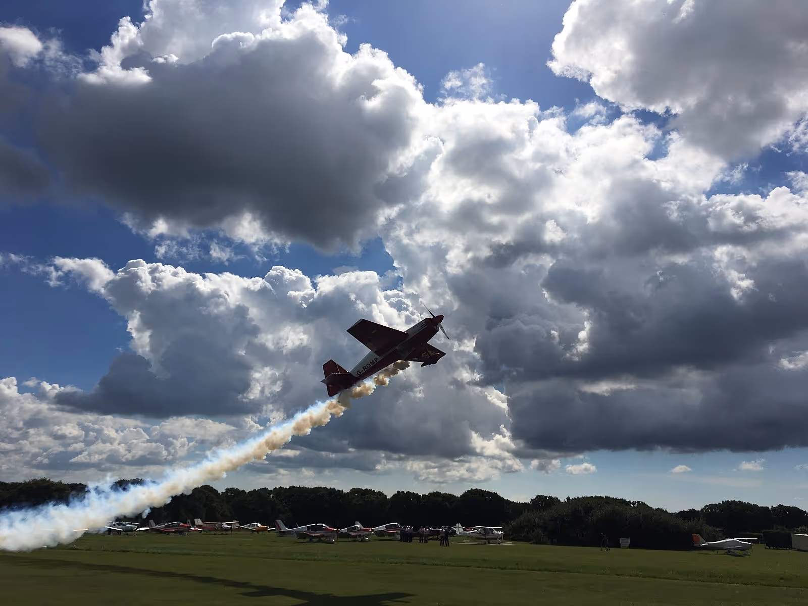 A red airplane performs an aerial stunt, leaving a trail of smoke against a cloudy sky.