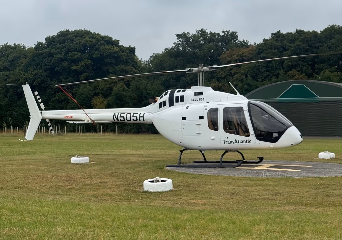 A white helicopter labeled “TransAtlantic” is parked on a grassy helipad.