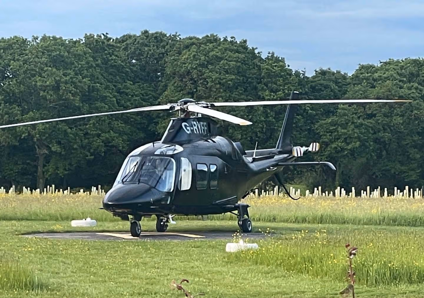 A black helicopter is parked on a grassy field with trees in the background.