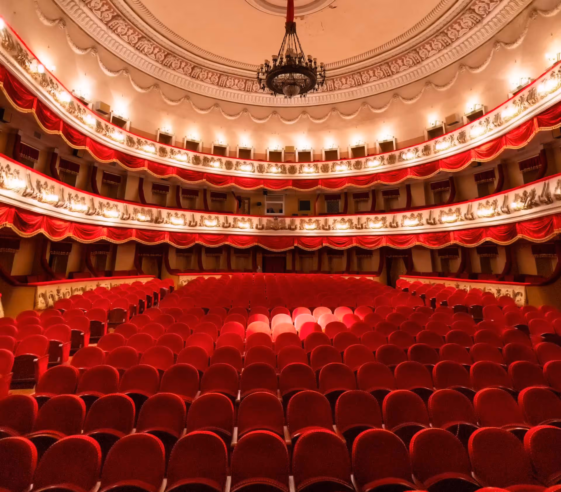 A spacious theater with rows of red seats facing an elaborate stage and ceiling chandelier.