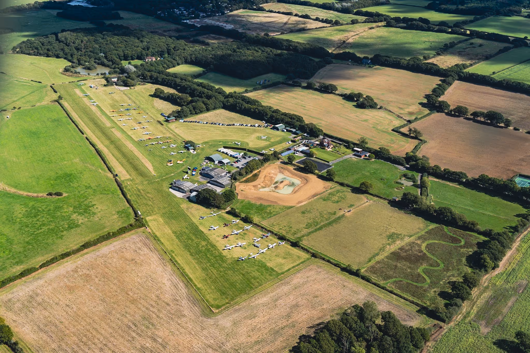 A small airfield with several parked airplanes surrounded by green fields, viewed from above.