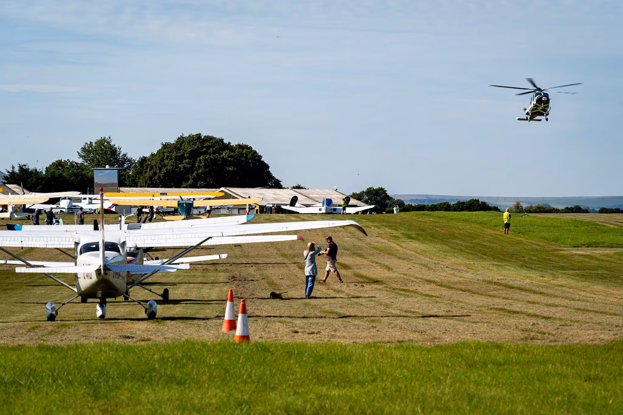 A person directs a small airplane on a grassy field, while a helicopter flies nearby.