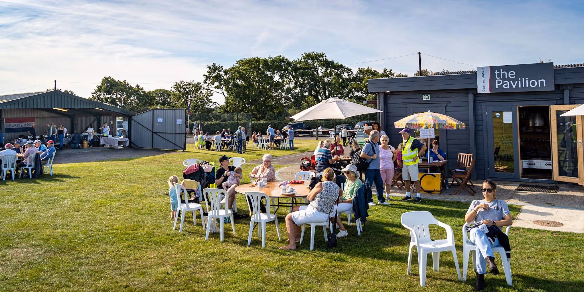 People sitting at outdoor tables eating, with a food stall and pavilion nearby under a sunny sky.