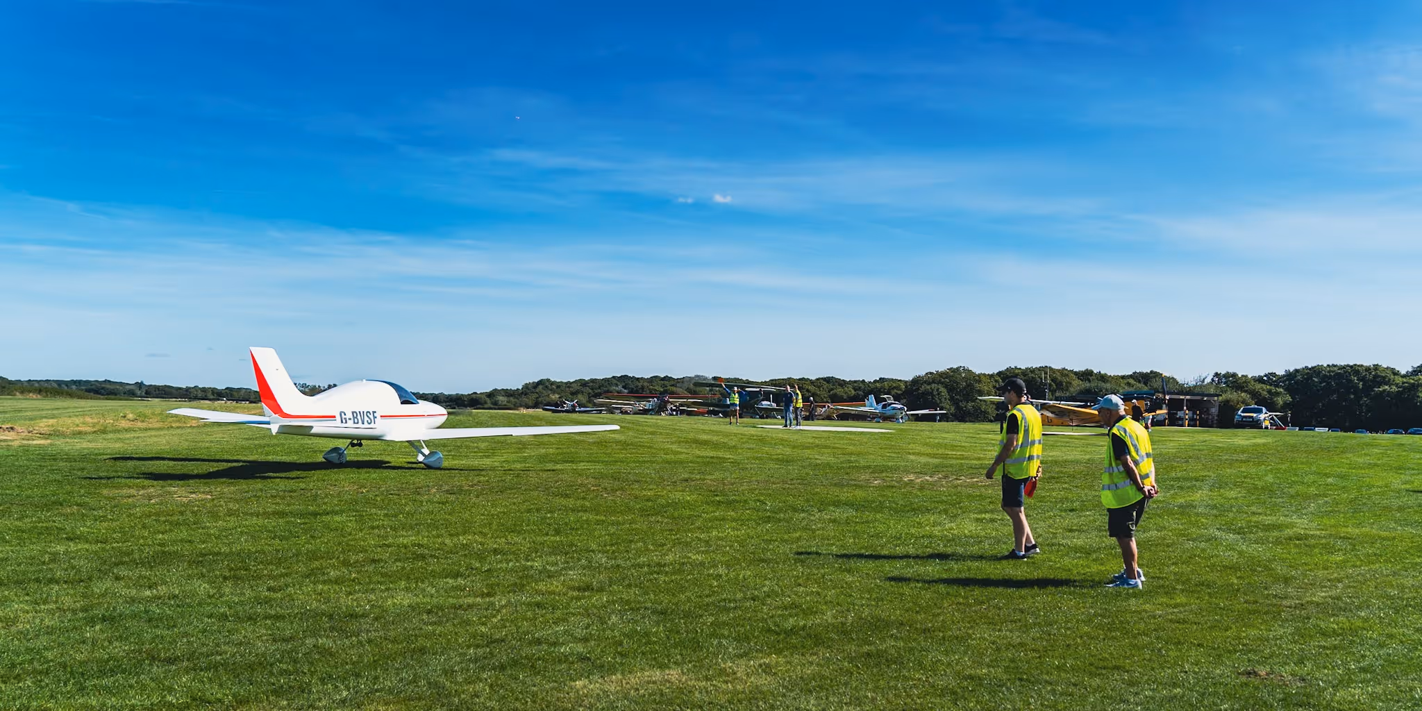 Two people in yellow vests watch a small airplane on a grassy field.