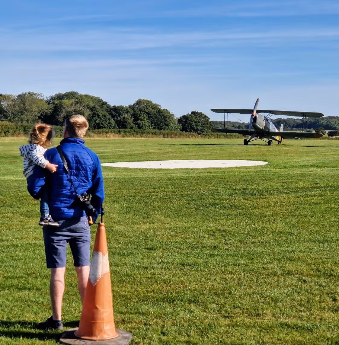 A man holding a child watches a biplane taxiing on a grassy field.
