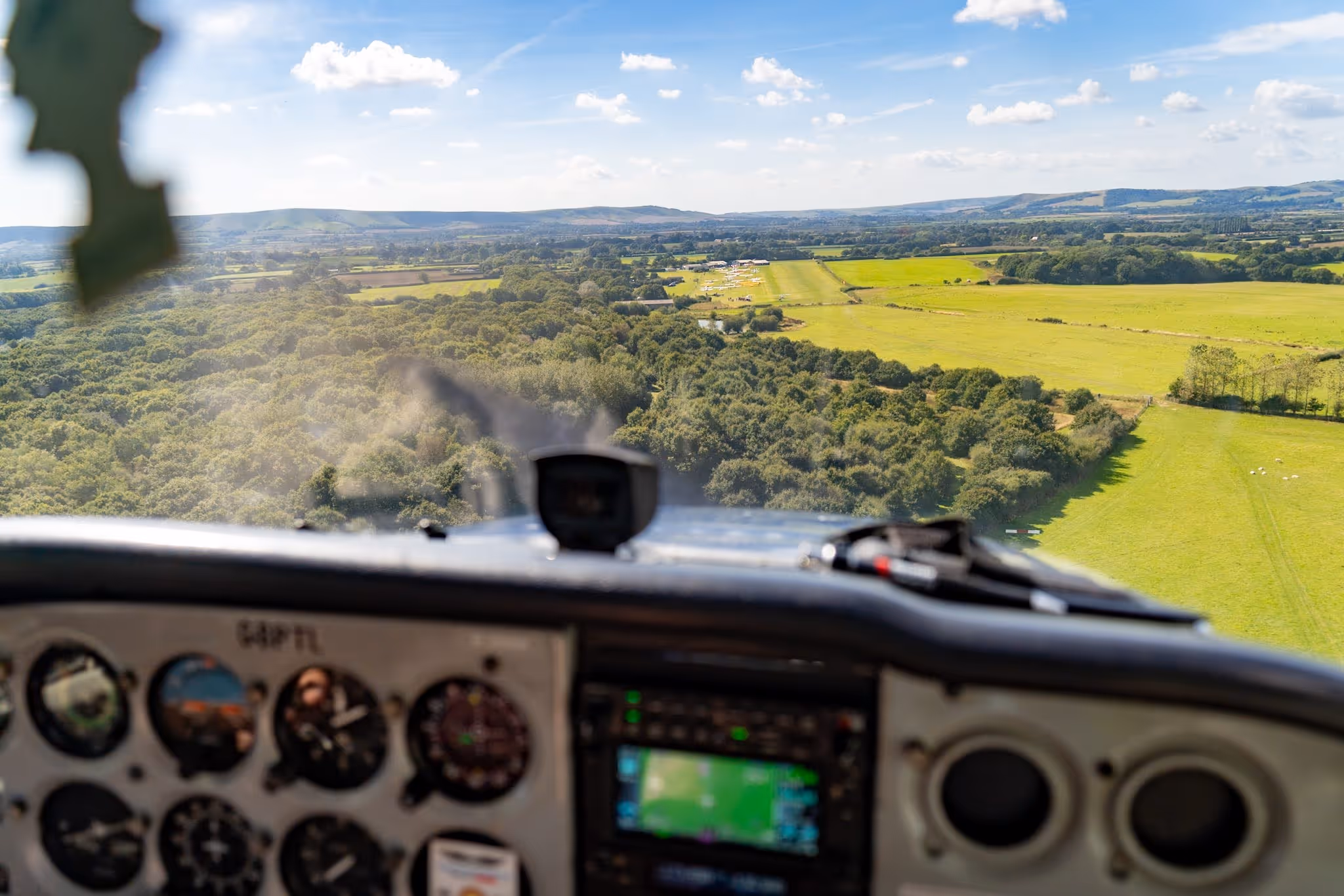 View from inside small airplane cockpit flying over green fields and trees under a clear blue sky.