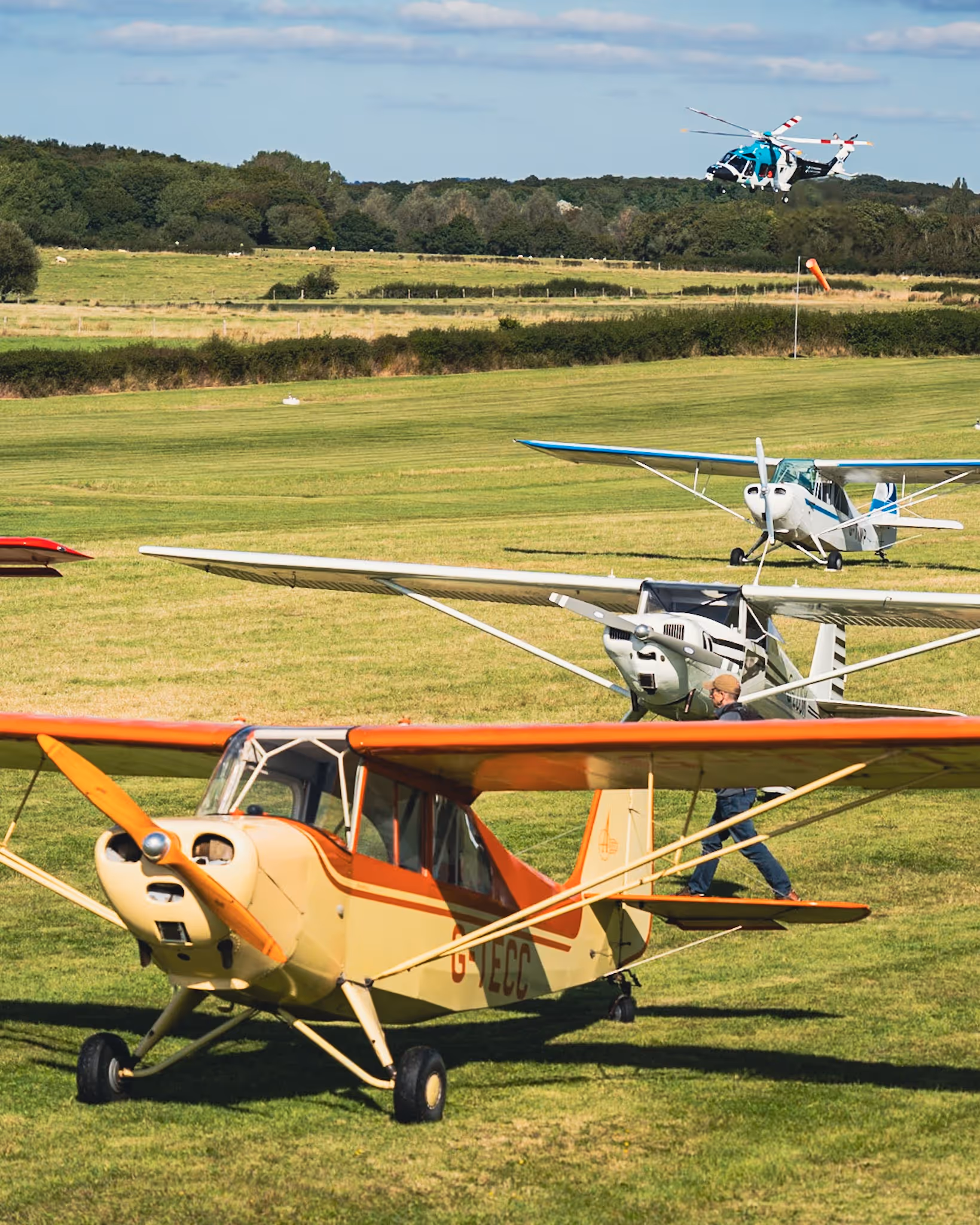 A person walks beside a small yellow and orange airplane on a grassy airfield with helicopters above.