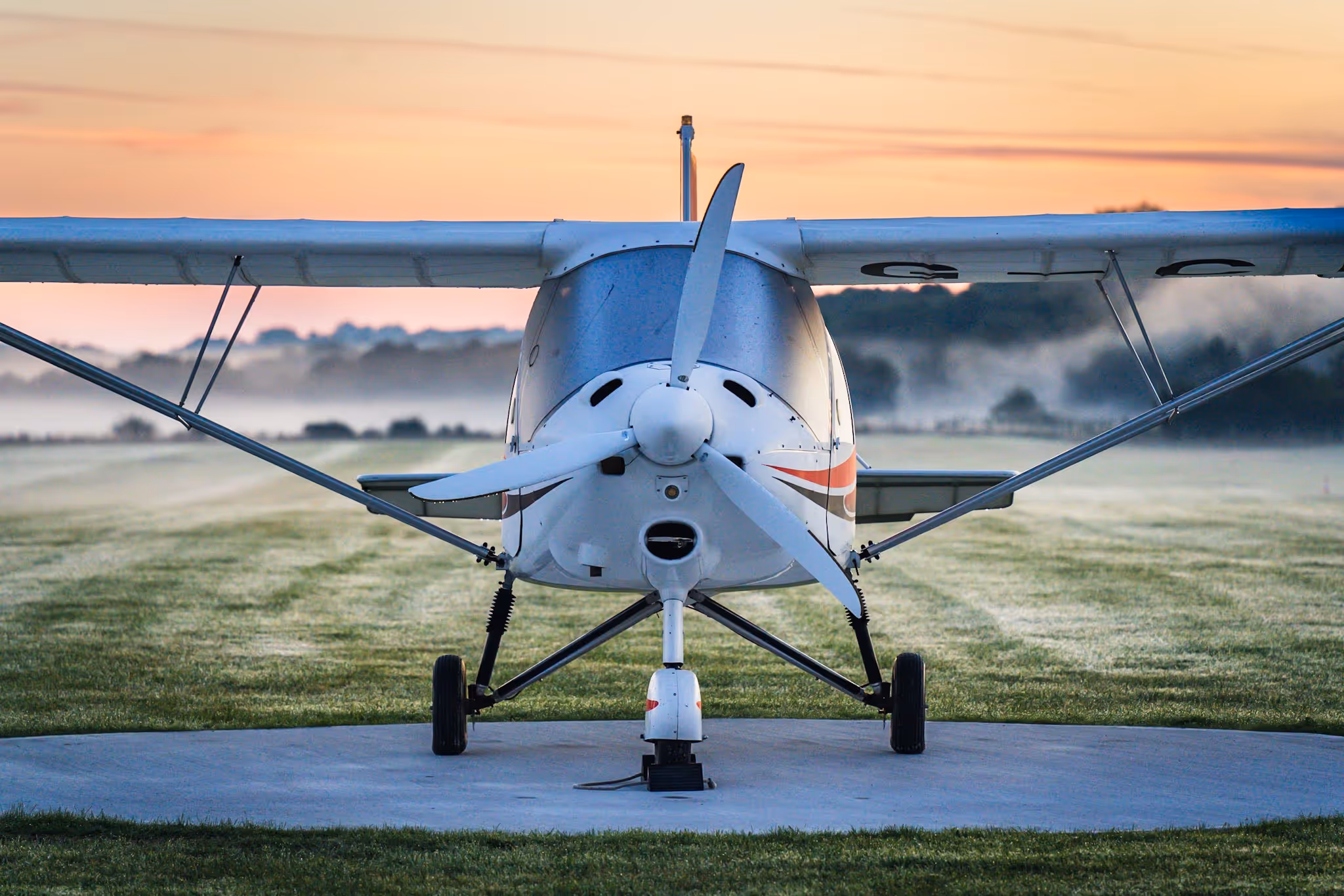 Small white propeller plane parked on grass runway at sunrise, facing forward.