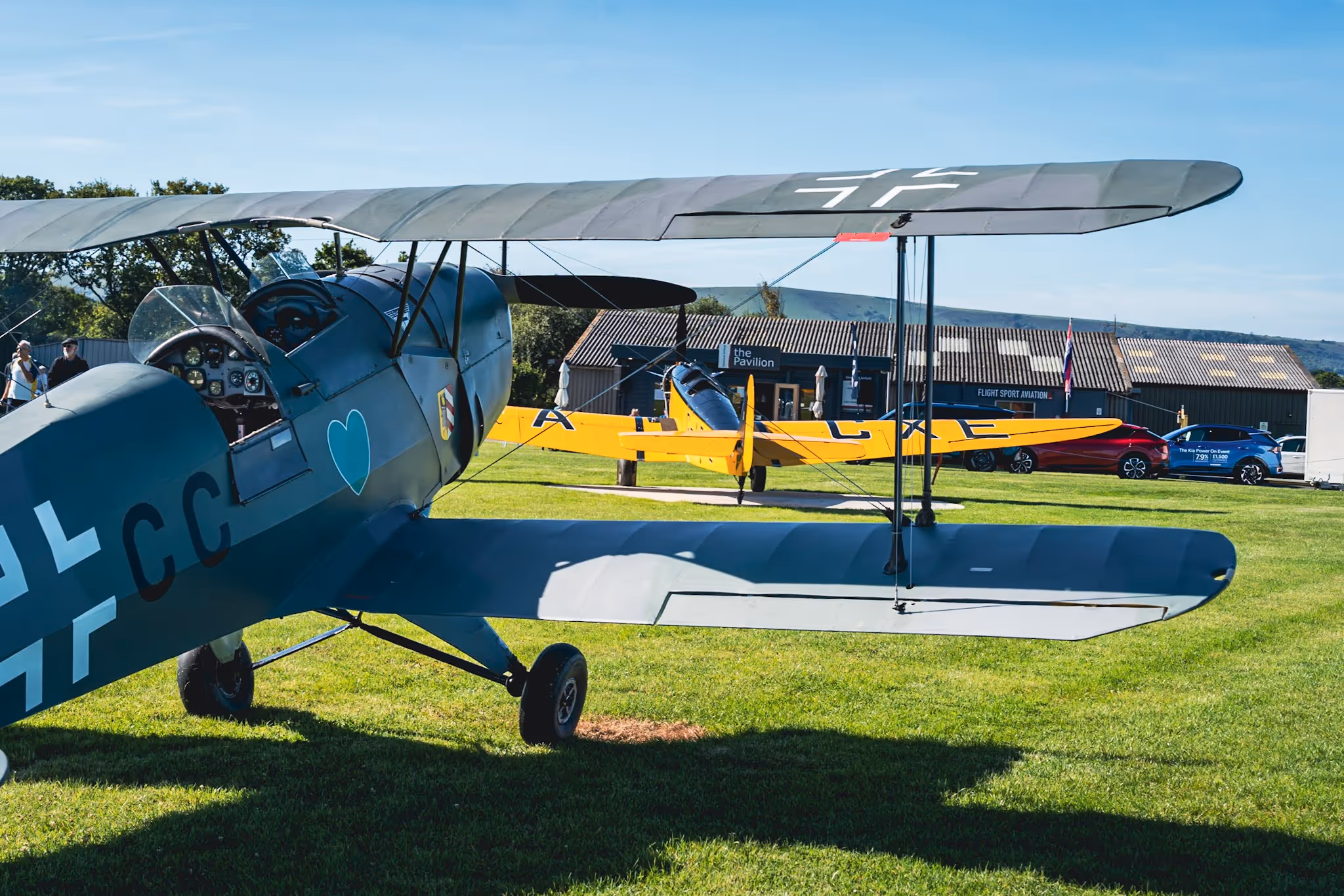 Two vintage biplanes are parked on a grassy airfield with a hangar in the background.