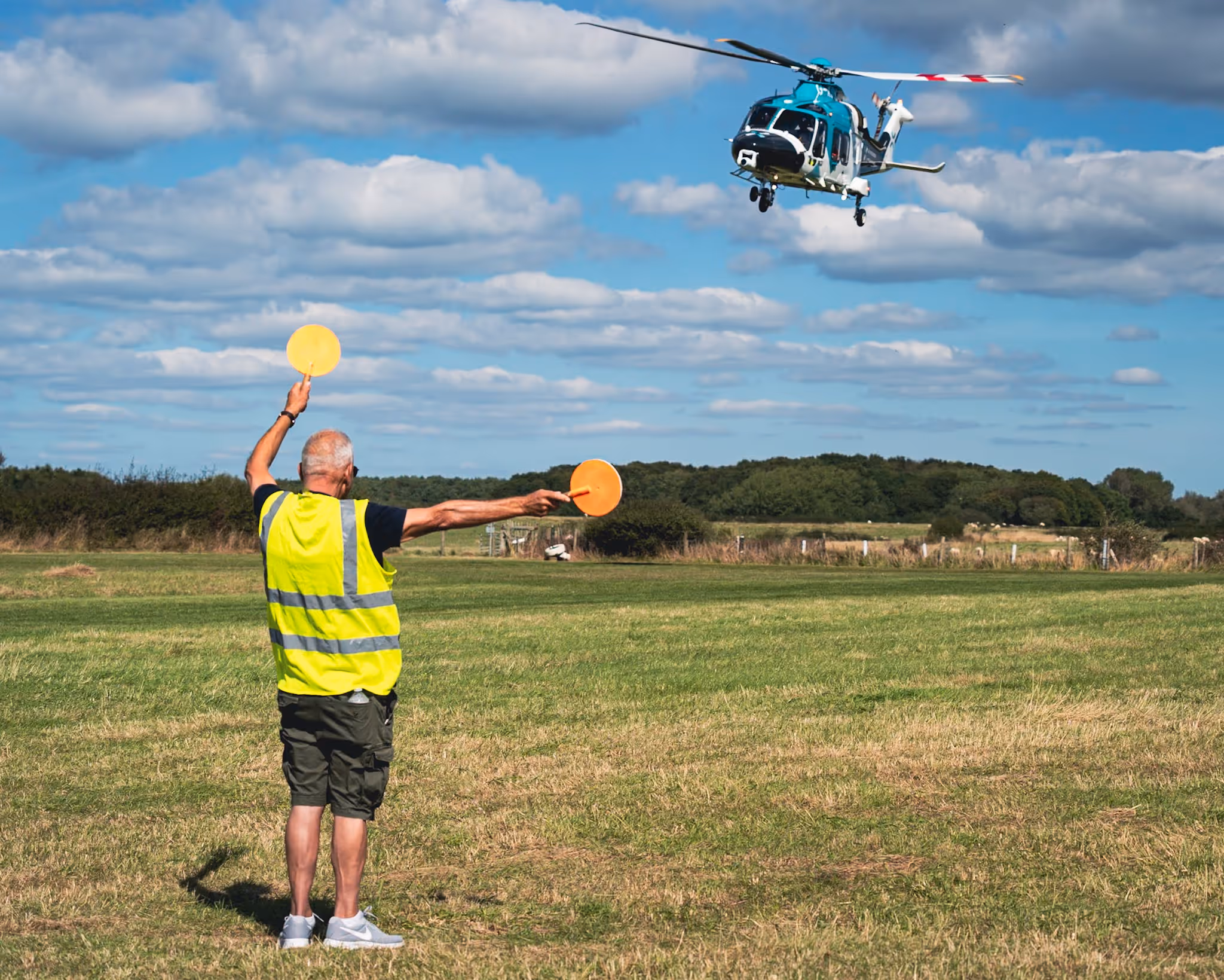 A man in a yellow vest guides a helicopter landing in an open grassy field.
