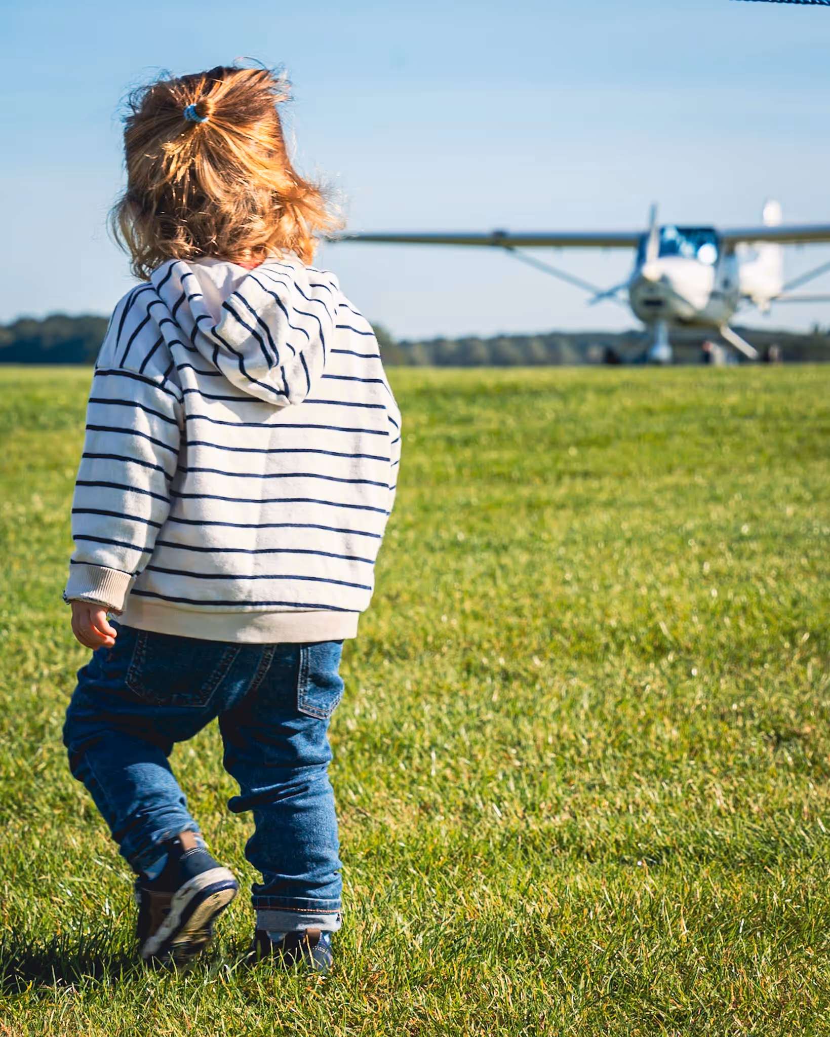 Child in striped hoodie walks on grass toward a small airplane on a clear day.