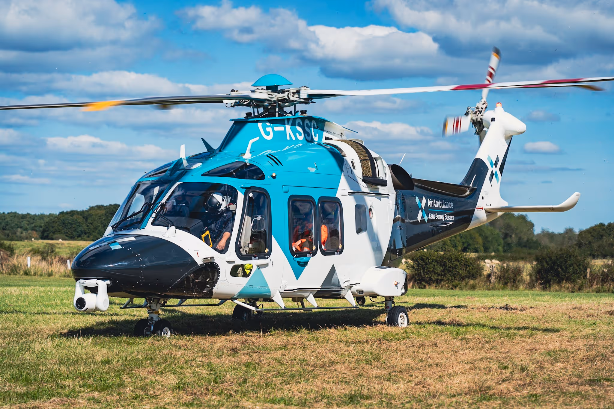 A teal and white helicopter lands on a grassy field under a blue sky with clouds.