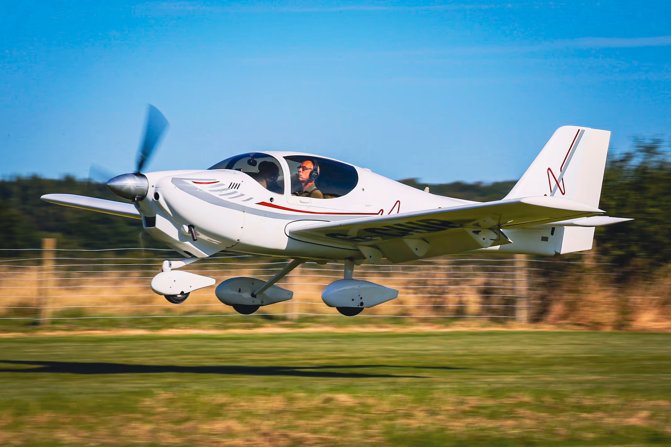 A small white plane flying low over a grassy field with a pilot inside.