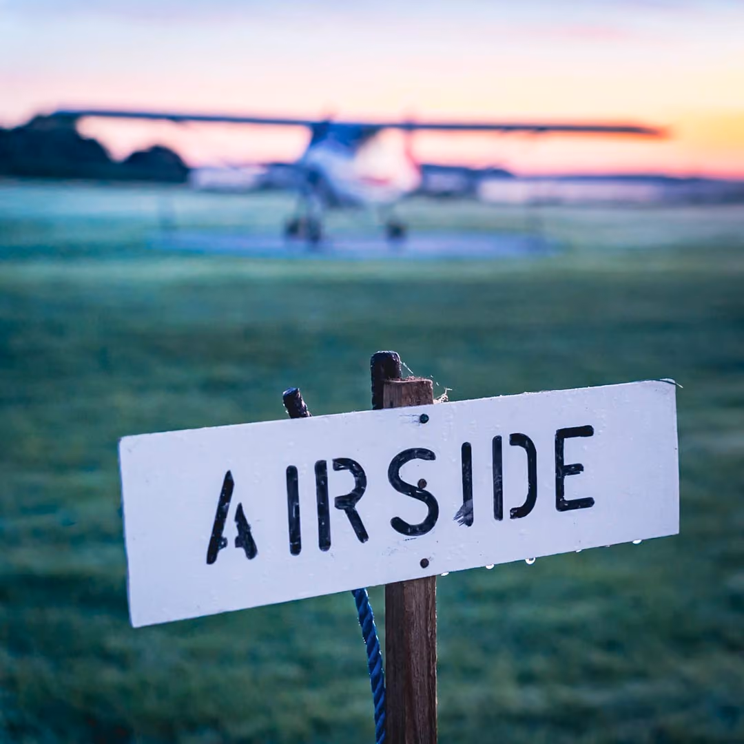 Sign with "Airside" in foreground, small plane on grass in background during sunrise.