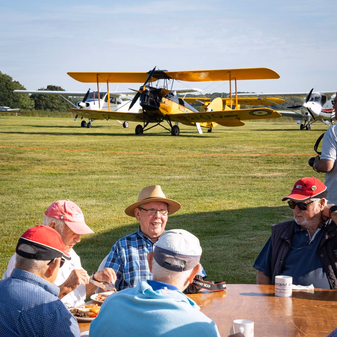 A group of older men are sitting at a table outdoors, eating, with planes nearby on the grass.