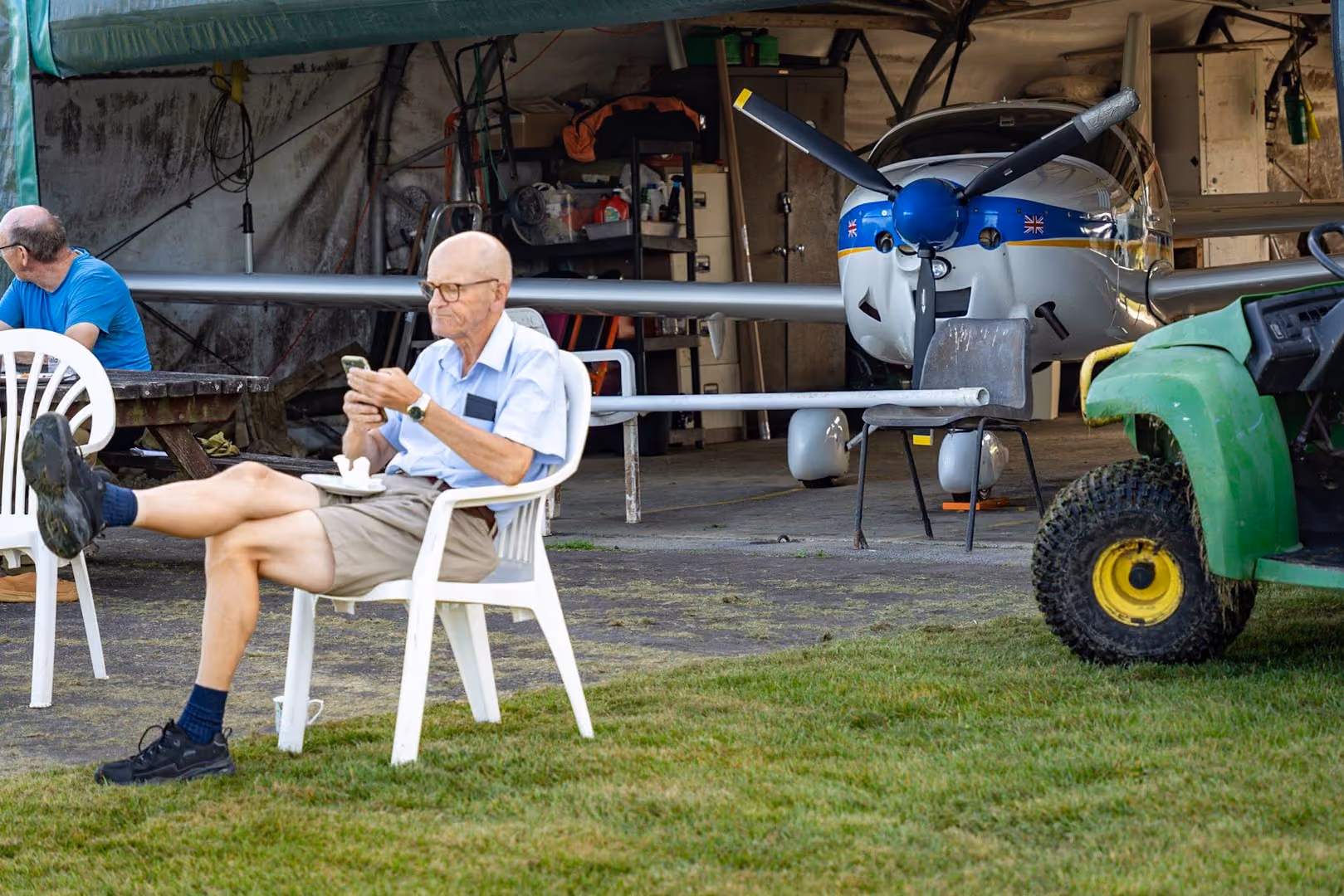 An older man sits on a chair checking his phone near a small aircraft in a hangar.