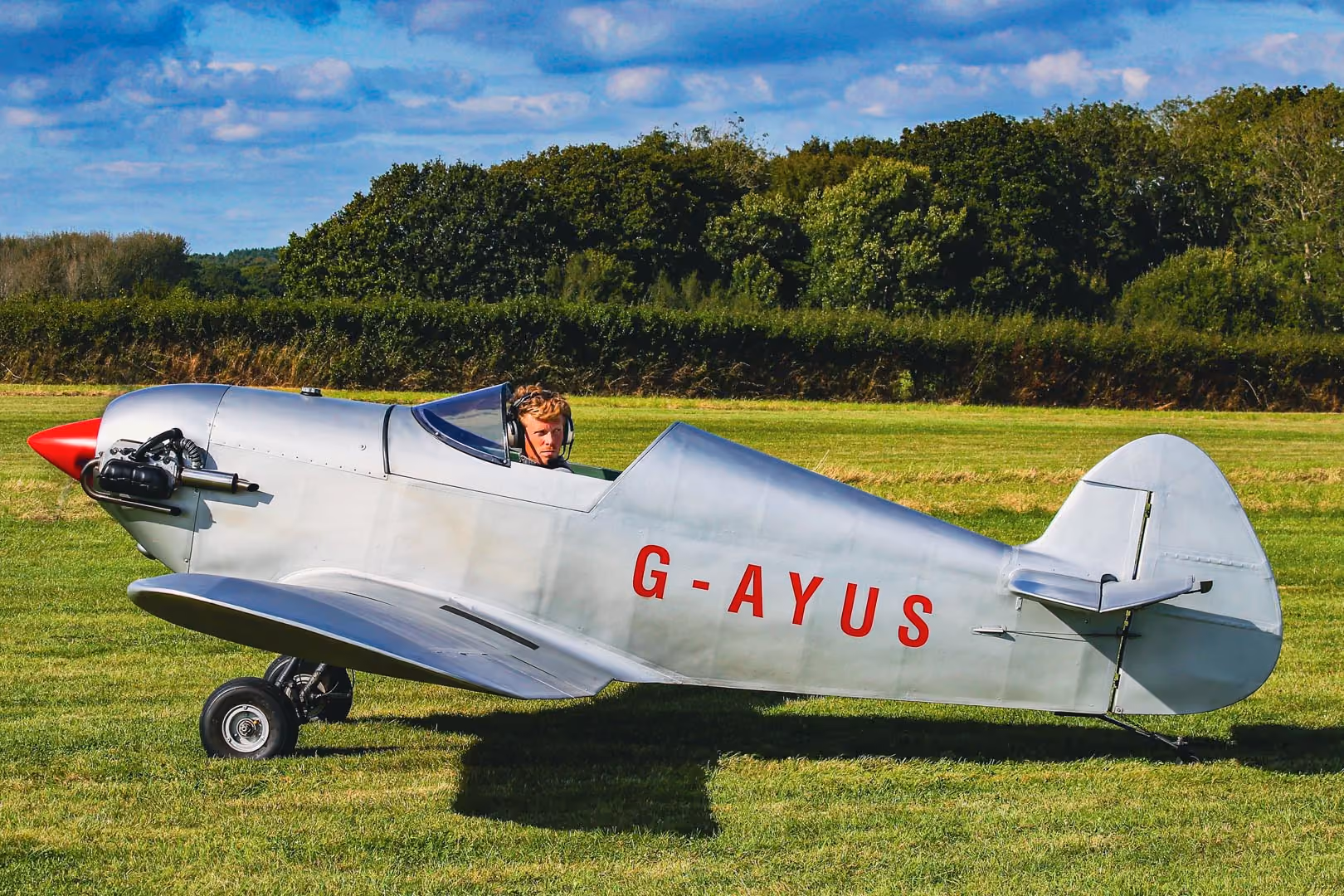 A small silver plane with a red nose stands on grass, with a person in the cockpit.