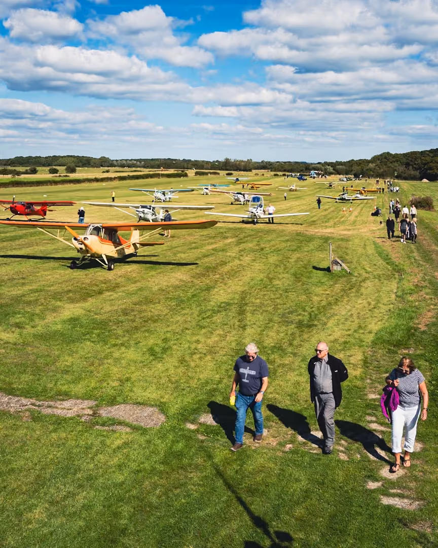 People walking on grass near parked small airplanes under a blue sky.