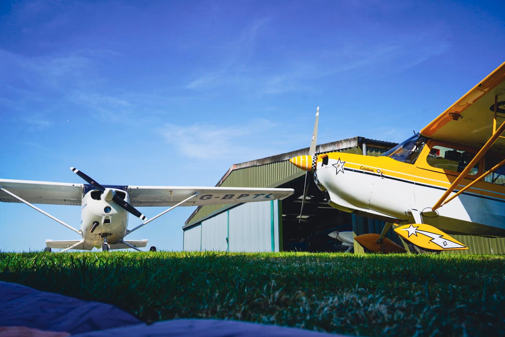 Two small airplanes parked on grass near a green hangar under a clear blue sky.