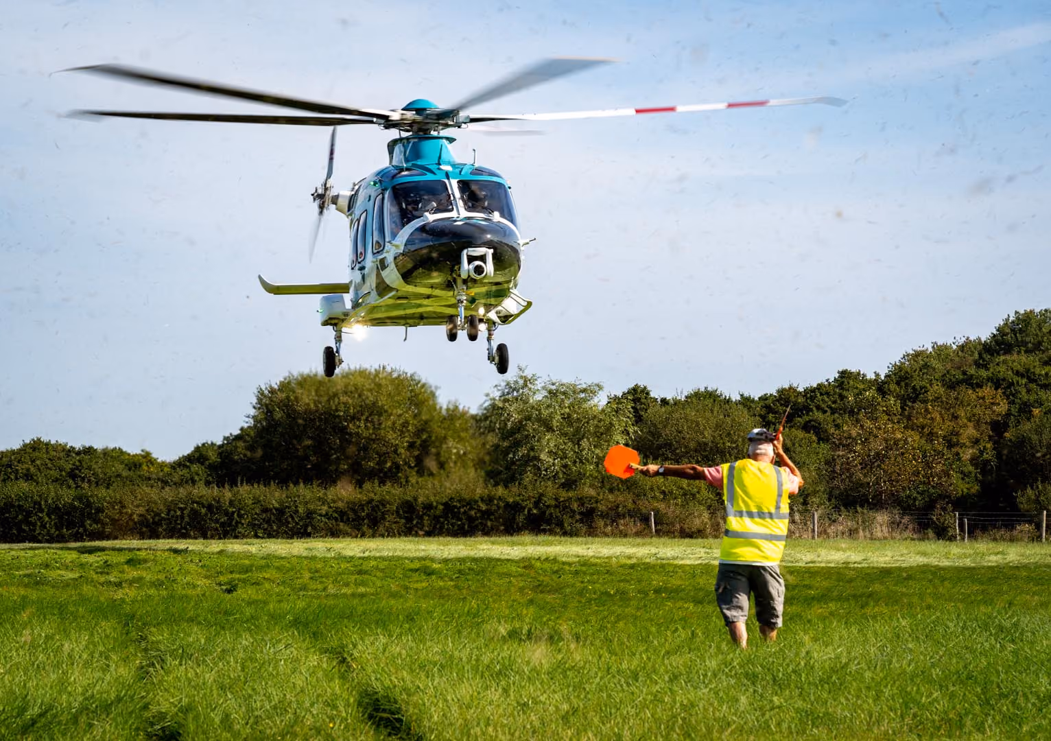 A person guides a helicopter landing in a grassy field using orange signals.